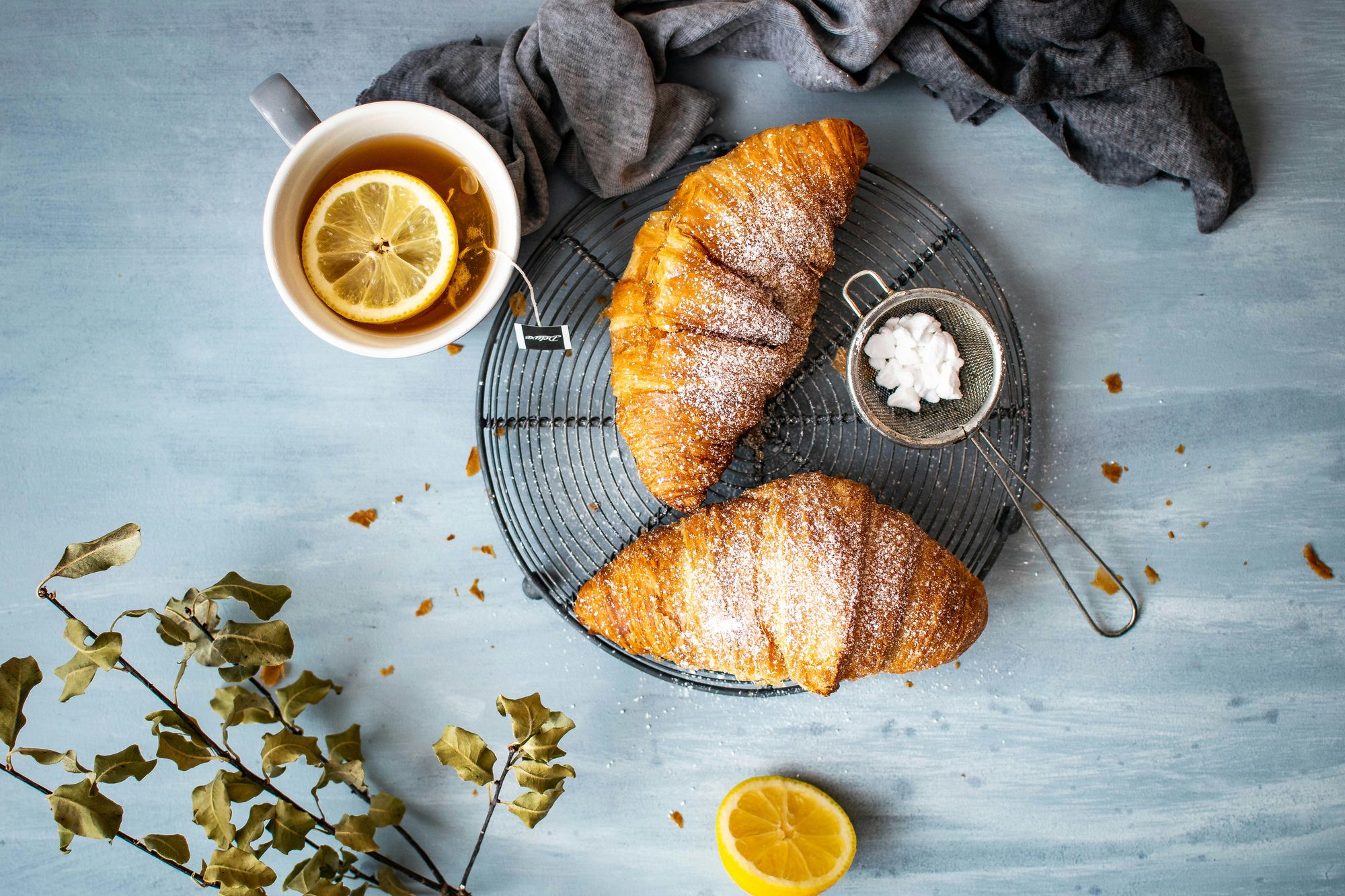 Zwei Croissants mit Puderzucker auf einem Gitterrost, dazu Tee mit Zitrone